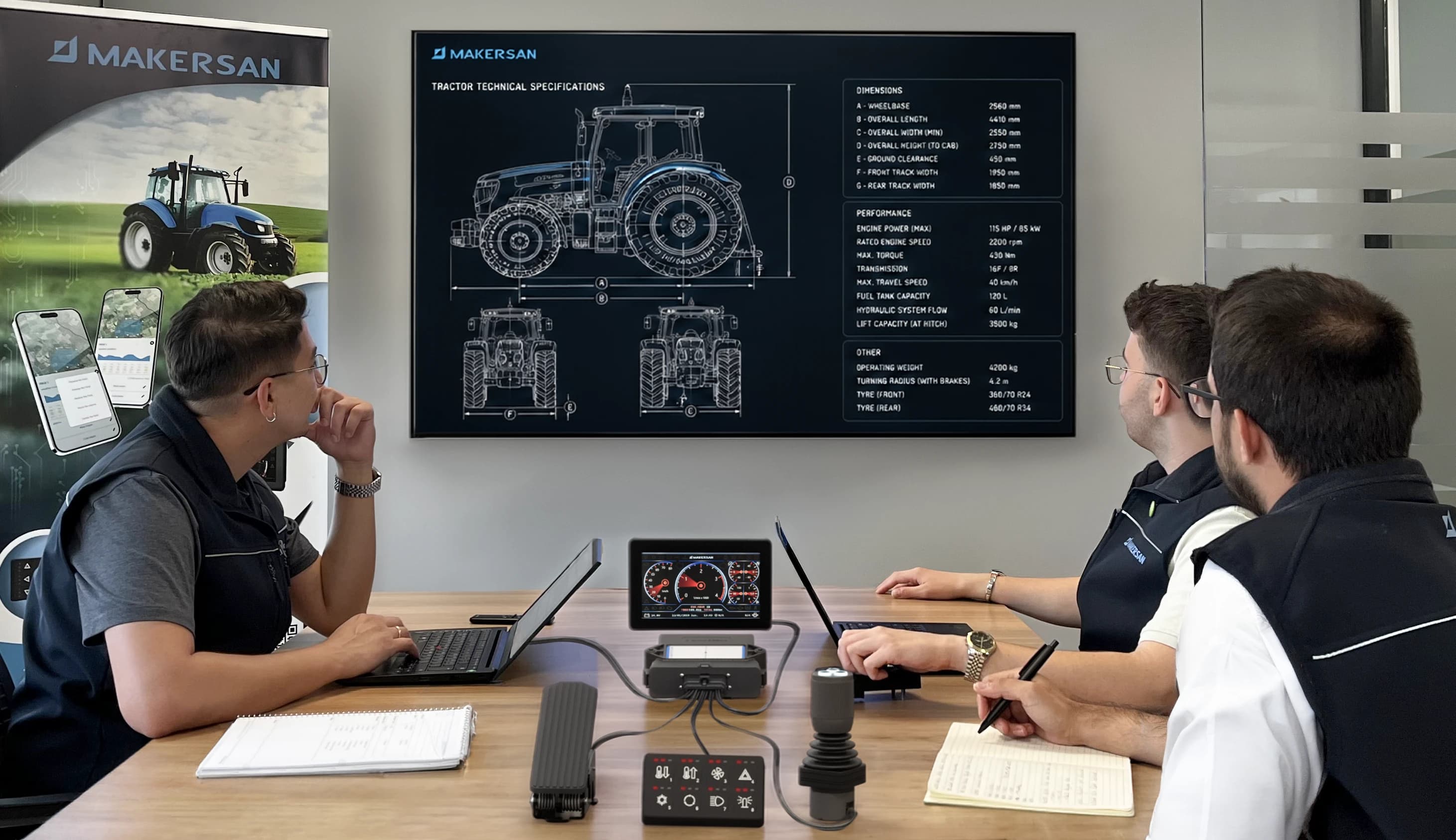 Three Makersan engineers reviewing a pedal assembly on a wall display, with the physical pedals, grips and sensors on the table in front of them